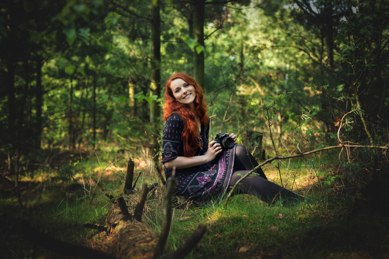 rothaarige Frau mit Kamera in der Hand sitzt auf einem Baumstamm